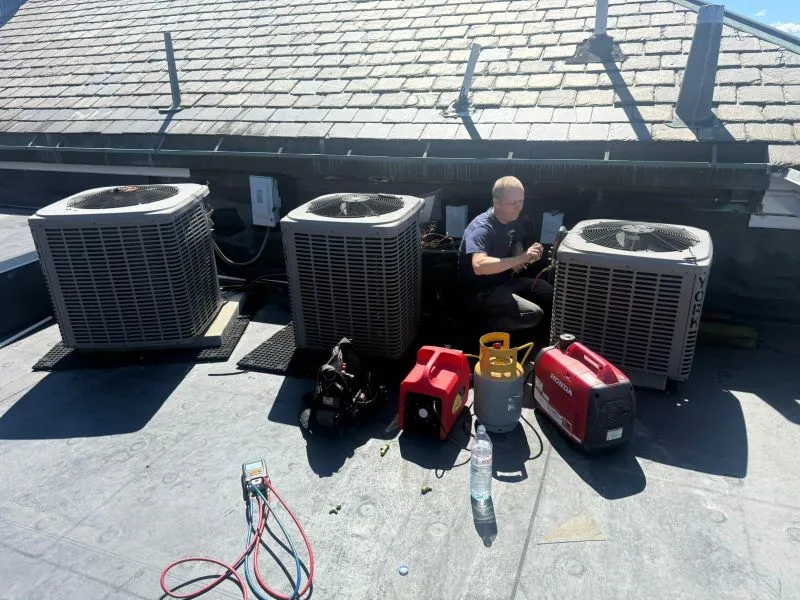 oltrom-ac1 HVAC technician on a flat rooftop servicing three outdoor AC units with tools and equipment nearby, sunny day.