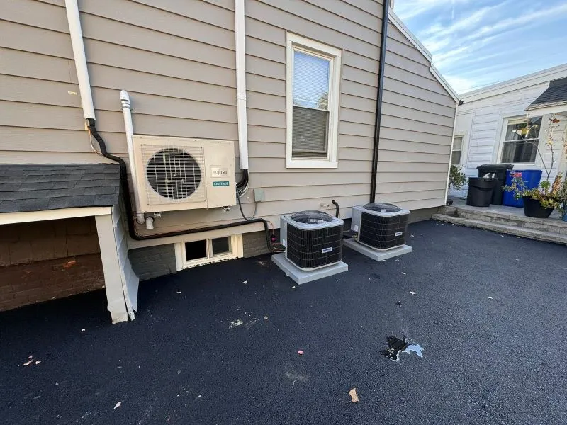 OLTROM installation AC in Garfield NJ for AIM Exterior view of a house showing two outdoor AC condensers on concrete pads beside a wall-mounted unit and a basement vent. 2. Back yard area with two air conditioning condensers on concrete pads and a wall-mounted outdoor unit, near a window. 3. Two outdoor AC units on a flat asphalt surface beside a gray house wall, with a window and utility pipes. 4. HVAC equipment outside a home: wall-mounted unit and two condensers on elevated pads. 5. Side of house showing split-system outdoor unit and two condensing units on concrete bases.