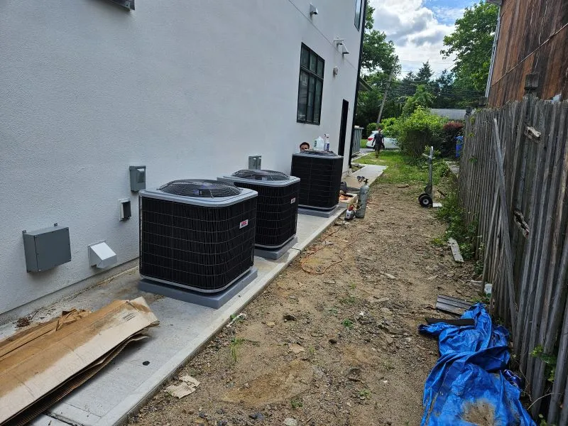 oltrom-ac2 Three outdoor air conditioning units sit on concrete pads along the white building wall, with a dirt path, fence, and construction materials nearby.