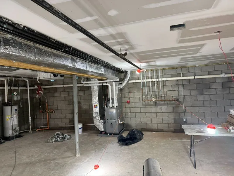 oltrom-ac8 Basement under renovation with exposed HVAC ducts, a furnace, water heater, and plumbing along concrete block walls; tools and a folding table are nearby.