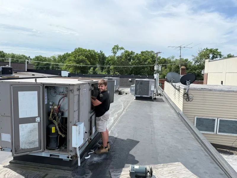 Technician on a flat rooftop servicing an outdoor HVAC Technician on a flat rooftop servicing an outdoor HVAC unit, with wiring exposed inside the open panel of the unit.